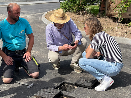 Zwei Ingenieur:innen und ein Mitglied des Bauteams knien auf dem Asphalt über der Öffnung zum Kugelventil.  und ein Mitglied des Bauti